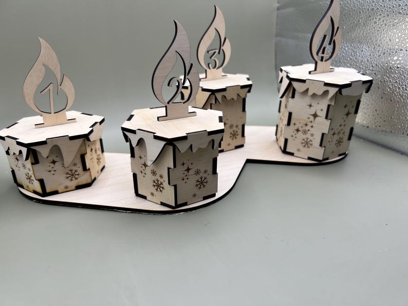 May include: Four wooden candle holders with a snowflake design. Each holder has a number on it, 1, 2, 3, and 4. The holders are arranged in a semi-circle on a wooden base.