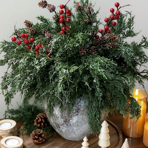 May include: A round, textured gray ceramic vase filled with lush green evergreen branches, small brown pinecones, and bright red berries. The arrangement is displayed alongside small wooden tree figurines and candles.