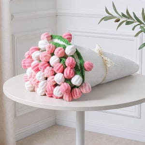 May include: A bouquet of crocheted flowers in shades of pink and white, wrapped in a white cone-shaped holder. The flowers are arranged with a green crocheted stem and a cream-colored ribbon tied around the holder.