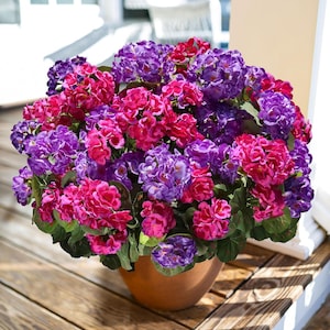 May include: A terracotta pot overflowing with vibrant, artificial hydrangeas. The flowers are a mix of deep purple, fuchsia, and red hues, with green foliage. The pot sits on a wooden deck, creating a colorful floral display.