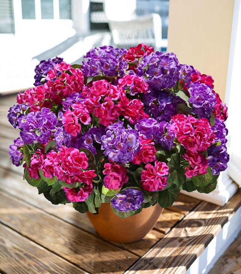 May include: A terracotta pot overflowing with vibrant, artificial hydrangeas. The flowers are a mix of deep purple, fuchsia, and red hues, with green foliage. The pot sits on a wooden deck, creating a colorful floral display.