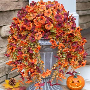 May include: A decorative urn filled with an arrangement of artificial flowers in shades of orange, red, and burgundy. The flowers feature small, five-petaled blooms and green foliage that cascades over the sides of the gray, fluted pedestal container.