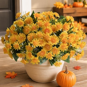 May include: A large, rounded arrangement of vibrant yellow artificial chrysanthemum flowers with green foliage, housed in a simple, matte beige bowl-shaped planter. A single orange pumpkin and several scattered orange maple leaves sit nearby.