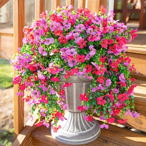 May include: A gray urn overflowing with vibrant pink and purple flowers and green foliage. The urn is placed on a wooden deck. The flowers cascade over the sides of the urn, creating a lush, full appearance. The flowers are in full bloom.