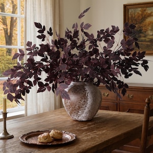 May include: A round, textured brown and white ceramic vase filled with dark purple, serrated-edged foliage and small berries. The vase sits on a wooden table next to a copper-toned metal tray holding two round, light-colored baked goods.