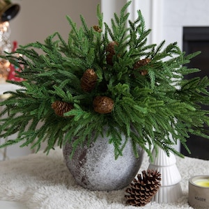 May include: A decorative arrangement featuring vibrant green faux evergreen branches and several brown pinecones. The arrangement is in a textured, gray ceramic pot. A white ceramic tree and a lit candle are also visible.