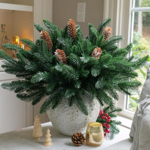 May include: A decorative arrangement featuring a large, textured, light gray vase filled with lush green faux pine branches and several brown pine cones. Additional wooden Christmas trees, a gold candle holder, and red berries complete the holiday display.
