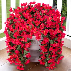 May include: A large, artificial hanging plant with bright red flowers and green leaves. The plant is in a white planter and is hanging over the edge of a wooden deck.