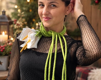 Può includere: Una sciarpa in feltro con fiori di calla bianchi con centri gialli e lunghi steli verde brillante che si avvolgono attorno al collo. La sciarpa è indossata sopra un top nero a maniche lunghe con uno strato in rete trasparente e puntinato.