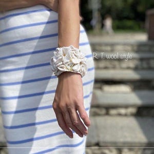 Puede incluir: Una pulsera de tela blanca con pétalos florales superpuestos y pequeños detalles en tono dorado. El texto R. T. wool style es visible en la esquina superior derecha. La pulsera se lleva en la muñeca con una falda de rayas blancas y azules.