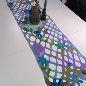 May include: A felt table runner in shades of purple, teal, and green, decorated with blue floral accents. The runner features a lattice design and is adorned with a vase of flowers and a candle holder.