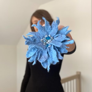 May include: A large, light blue felt flower brooch with a sparkling jewel center. The brooch has multiple layers of petals and is held up in front of a person wearing a black top. The flower is a striking accessory.