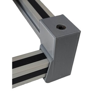 May include: A gray corner bracket connecting two metal rails. The rails are light gray with two black stripes. The bracket has a hole for a screw or bolt. The items are likely for a shelving or framing system.