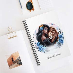 Peut inclure: Un carnet à spirales avec une couverture blanche illustrant un cercle représentant une femme aux longs cheveux noirs portant une couronne et un lion. La femme tient le lion et ils sont entourés de fleurs bleues. Le texte "Jésus est Sauveur" est imprimé sous l'illustration.