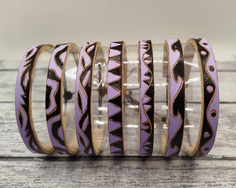 Può includere: Un set di sei braccialetti in pelle viola e nera con motivi geometrici. I braccialetti sono impilati uno sopra l'altro e hanno una finitura lucida.