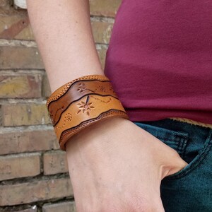 Könnte beinhalten: Ein braunes Lederarmband mit einem dekorativen Blumenmuster. Das Armband wird am Handgelenk getragen.