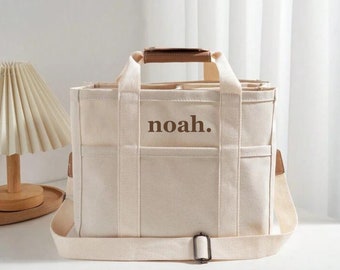 May include: A white canvas tote bag with brown leather handles and a brown leather strap. The bag has multiple pockets and the word "noah." is printed on the front in brown.