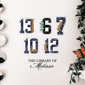 Op de afbeelding: Een witte muur met vijf blauwe en witte genummerde stickers met bloemenpatronen en gouden letters. De nummers zijn 13, 6, 7, 10 en 12. De tekst onder de nummers luidt "The Library of Melissa".