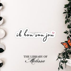 May include: A white wall with a sticker that reads "ik hou van jou" in black script with a pink heart. Below the sticker is the text "THE LIBRARY OF Melissa" in black script. Decorative hanging ceramic moon phases and butterflies.