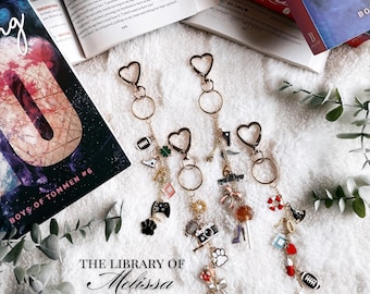 May include: Gold-toned keychains with heart-shaped clasps and various charms, including a paw print, camera, and high heel. The text "THE LIBRARY OF Melissa" is visible. Books are in the background.