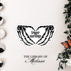 May include: A black and white sticker featuring a heart shape formed by skeleton hands. The words "trigger warnings" are in the center. The sticker is on a white wall, with the text "THE LIBRARY OF Melissa" below.