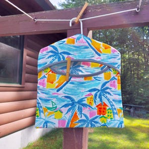May include: A colorful garment bag with a tropical print of palm trees, houses, and boats in shades of blue, yellow, orange, and pink. The bag is hanging on a white hanger and secured with wooden clothespins.