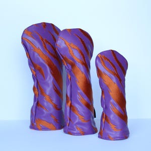 May include: Three golf club head covers in varying sizes. The covers are a vibrant purple with a bold, orange tiger stripe pattern. The covers are made of a smooth, possibly leather-like material.