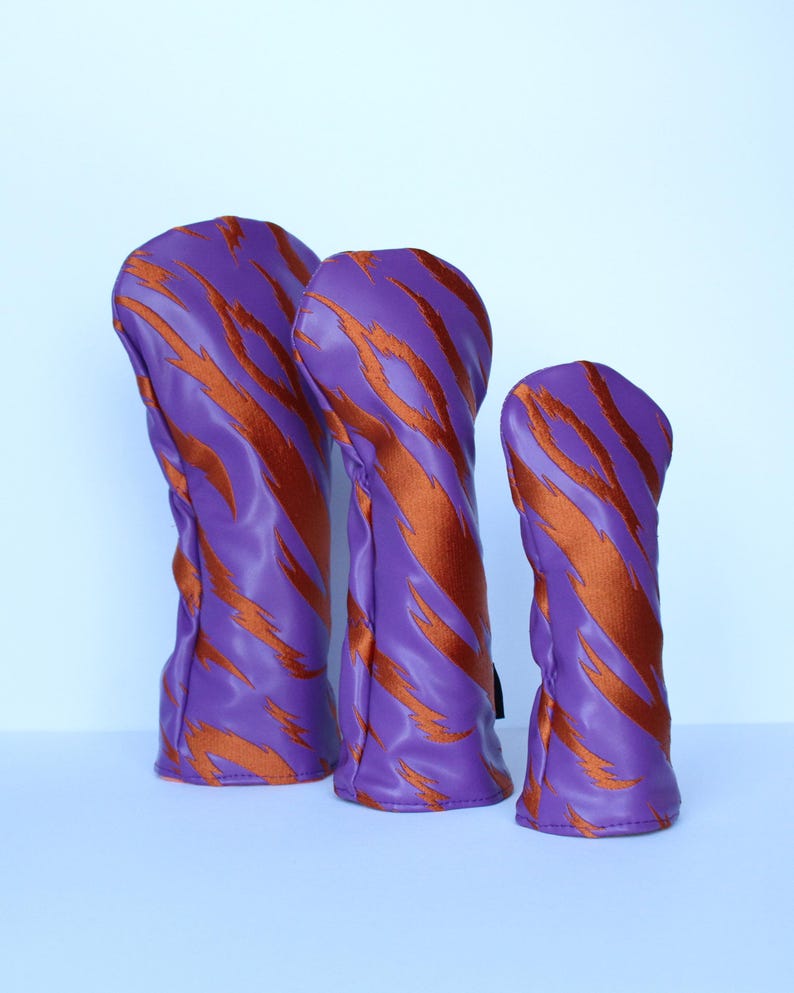 May include: Three golf club head covers in varying sizes. The covers are a vibrant purple with a bold, orange tiger stripe pattern. The covers are made of a smooth, possibly leather-like material.