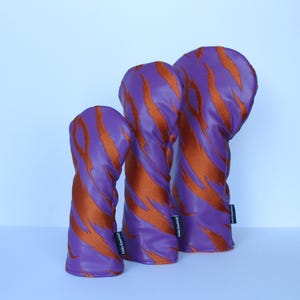 May include: Three golf club head covers in a row. The covers are purple with orange tiger stripe patterns. Each cover has a small black tag on the side. The covers are made of a shiny material.
