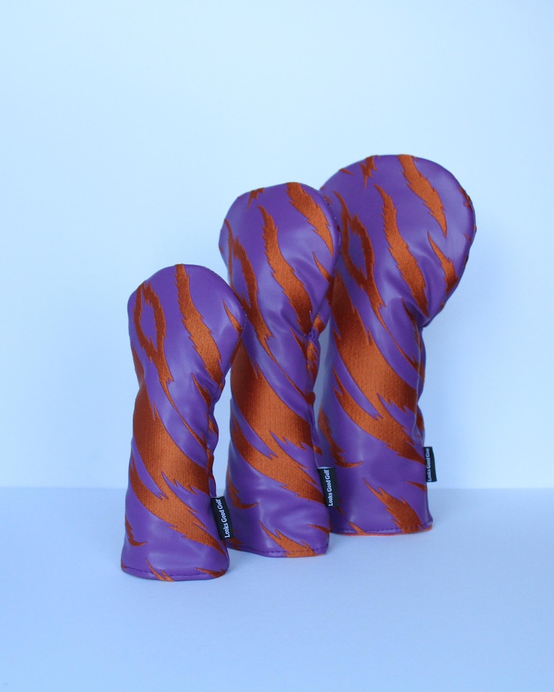 May include: Three golf club head covers in a row. The covers are purple with orange tiger stripe patterns. Each cover has a small black tag on the side. The covers are made of a shiny material.