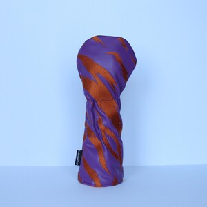 May include: Purple golf club head cover with a bold orange tiger stripe pattern. The cover has a stitched seam and a small black tag with white text. The golf club head cover is designed to protect golf clubs.