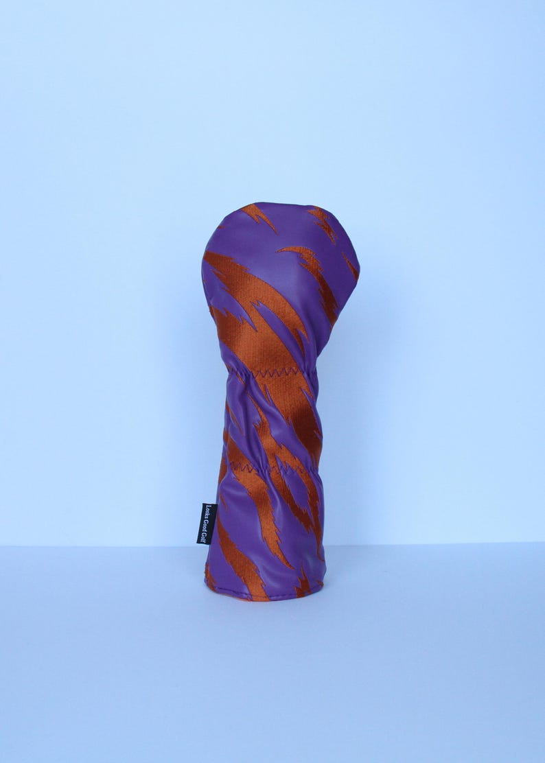 May include: Purple golf club head cover with a bold orange tiger stripe pattern. The cover has a stitched seam and a small black tag with white text. The golf club head cover is designed to protect golf clubs.