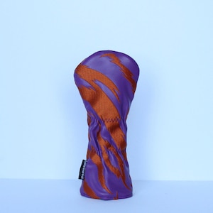 May include: Purple and orange golf club head cover with a dynamic, abstract design. The cover is made of a textured material and features a stitched seam. The label on the side reads "Looks Good Golf".