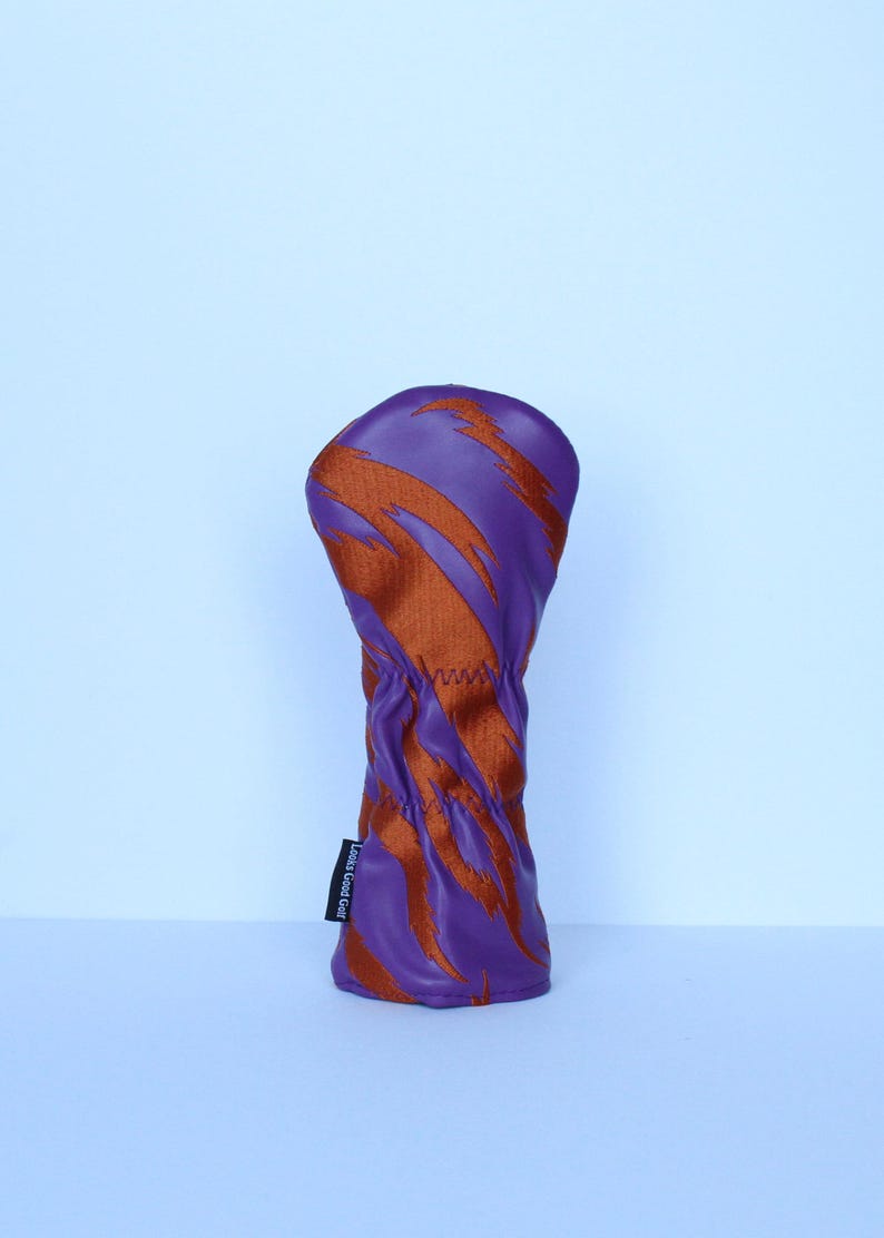 May include: Purple and orange golf club head cover with a dynamic, abstract design. The cover is made of a textured material and features a stitched seam. The label on the side reads "Looks Good Golf".