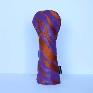 May include: Purple golf club head cover with orange tiger stripe design. The cover has a soft, textured appearance and a label that reads "Looks Good Golf". The cover is designed to protect golf clubs.