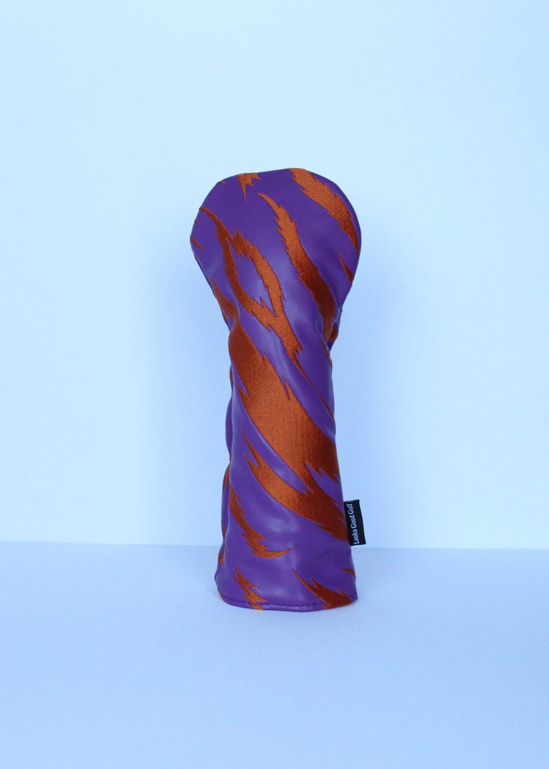 May include: Purple golf club head cover with orange tiger stripe design. The cover has a soft, textured appearance and a label that reads "Looks Good Golf". The cover is designed to protect golf clubs.