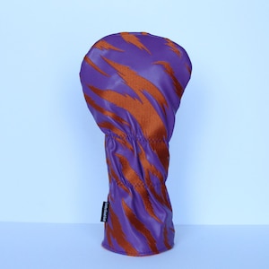 May include: A purple golf club head cover with an orange tiger stripe pattern. The cover is made of a smooth, synthetic material and has a stitched seam at the base. The label on the side reads "Looks Good Golf".