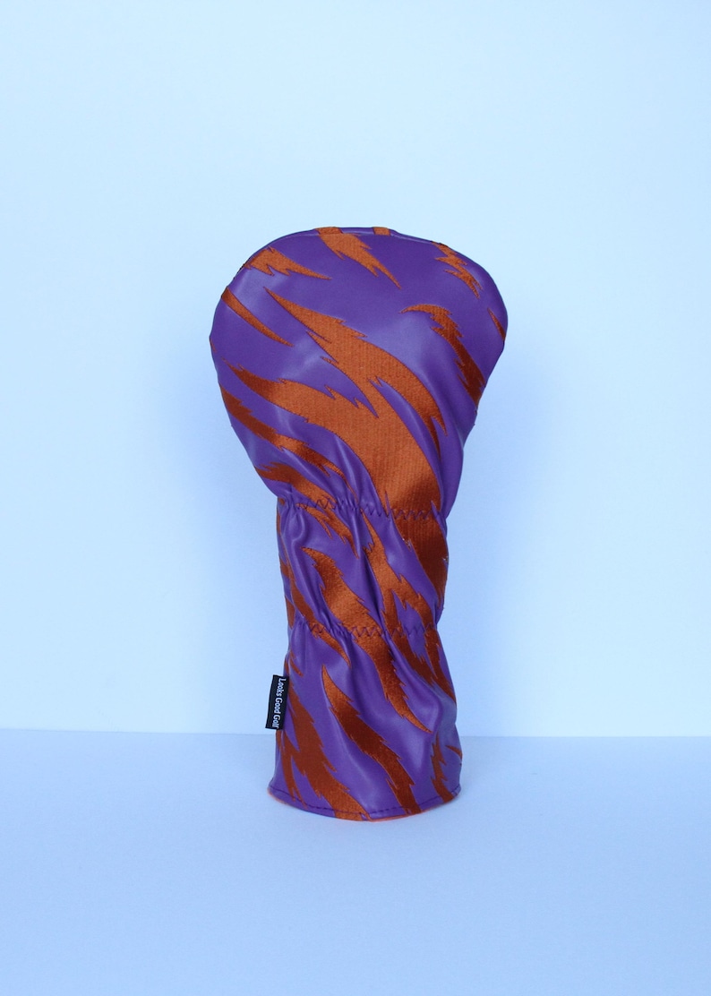 May include: A purple golf club head cover with an orange tiger stripe pattern. The cover is made of a smooth, synthetic material and has a stitched seam at the base. The label on the side reads "Looks Good Golf".