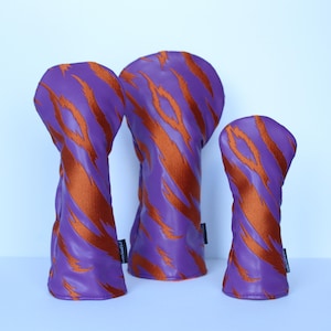 May include: Three golf club head covers in a row. The covers are purple with orange tiger stripe patterns. The covers are made of a leather-like material. The background is a light blue.