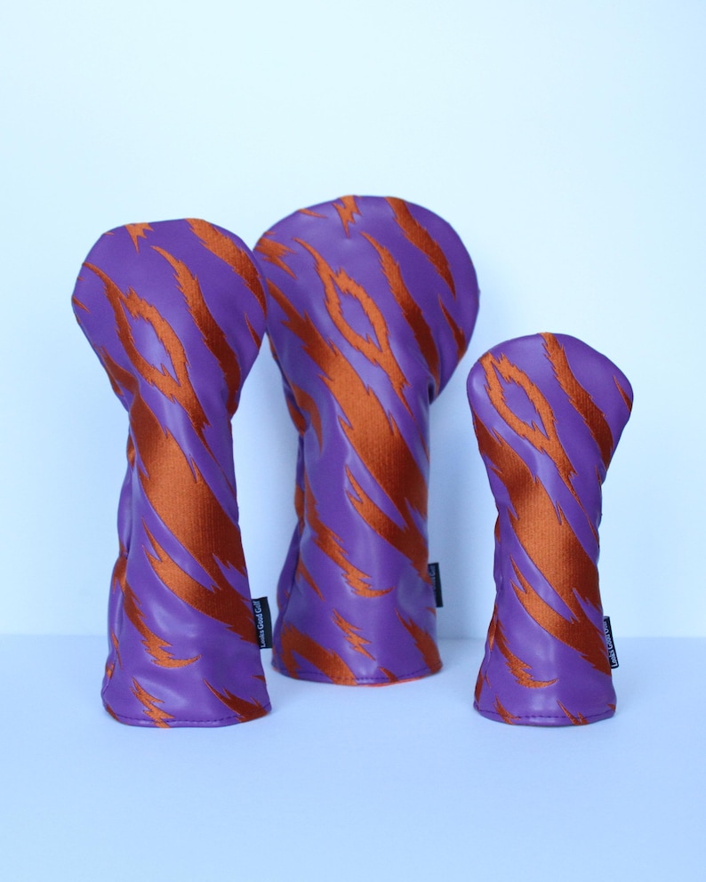 May include: Three golf club head covers in a row. The covers are purple with orange tiger stripe patterns. The covers are made of a leather-like material. The background is a light blue.