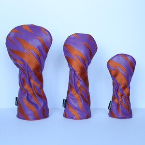 May include: Three golf club head covers in varying sizes. The covers are purple with orange tiger stripe patterns. The covers are made of a smooth material and have a stitched seam. Each cover has a small black tag.