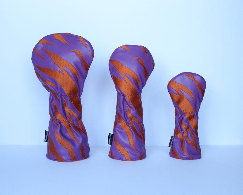 May include: Three golf club head covers in varying sizes. The covers are purple with orange tiger stripe patterns. The covers are made of a smooth material and have a stitched seam. Each cover has a small black tag.