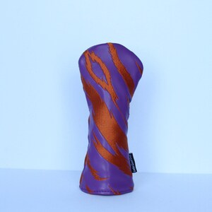 May include: A golf club head cover with a purple background and orange tiger stripe design. The cover has a cylindrical shape and a small black tag on the side with white text.