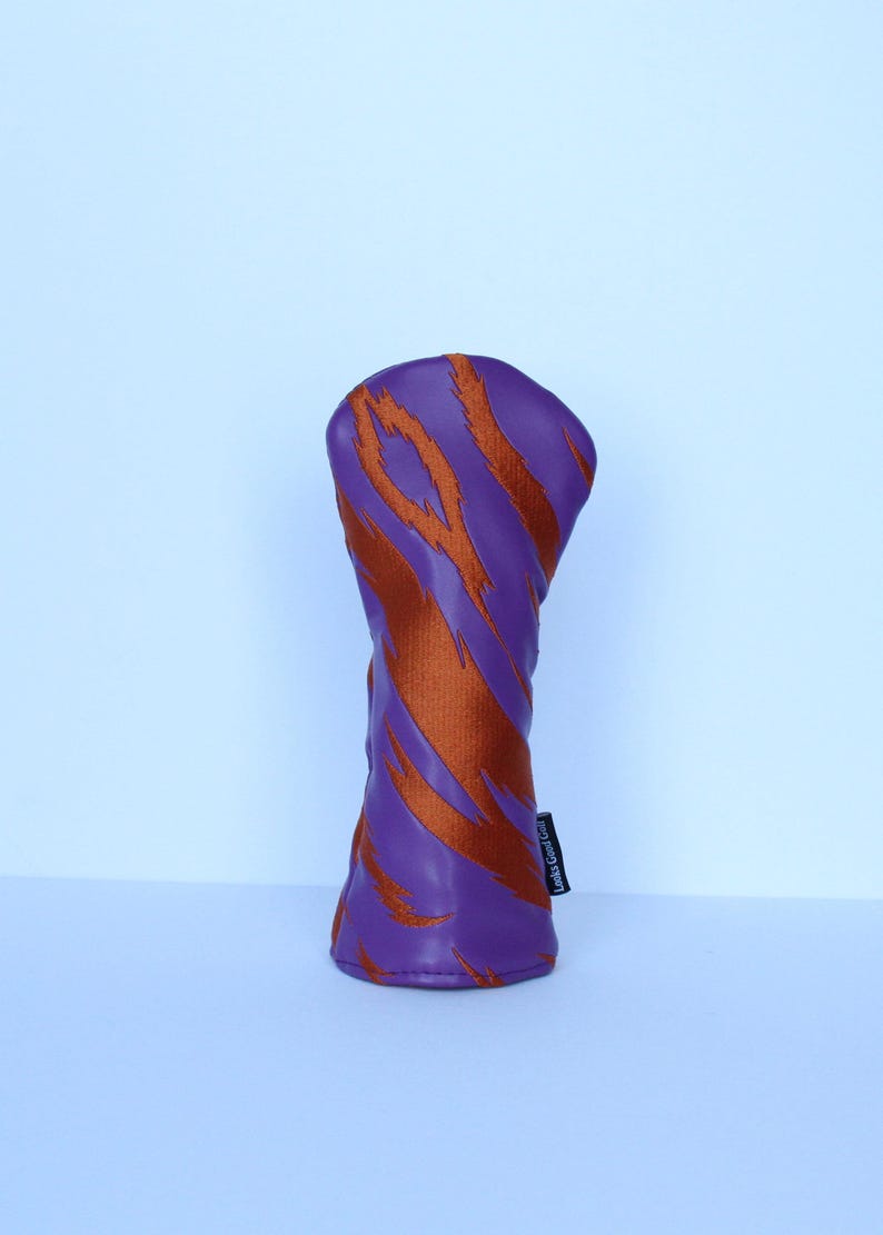 May include: A golf club head cover with a purple background and orange tiger stripe design. The cover has a cylindrical shape and a small black tag on the side with white text.