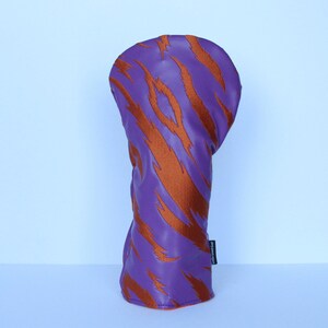 May include: Purple golf club head cover with orange tiger stripe design. The cover is made of a smooth material and has a rounded top. The cover is designed to protect the golf club head.