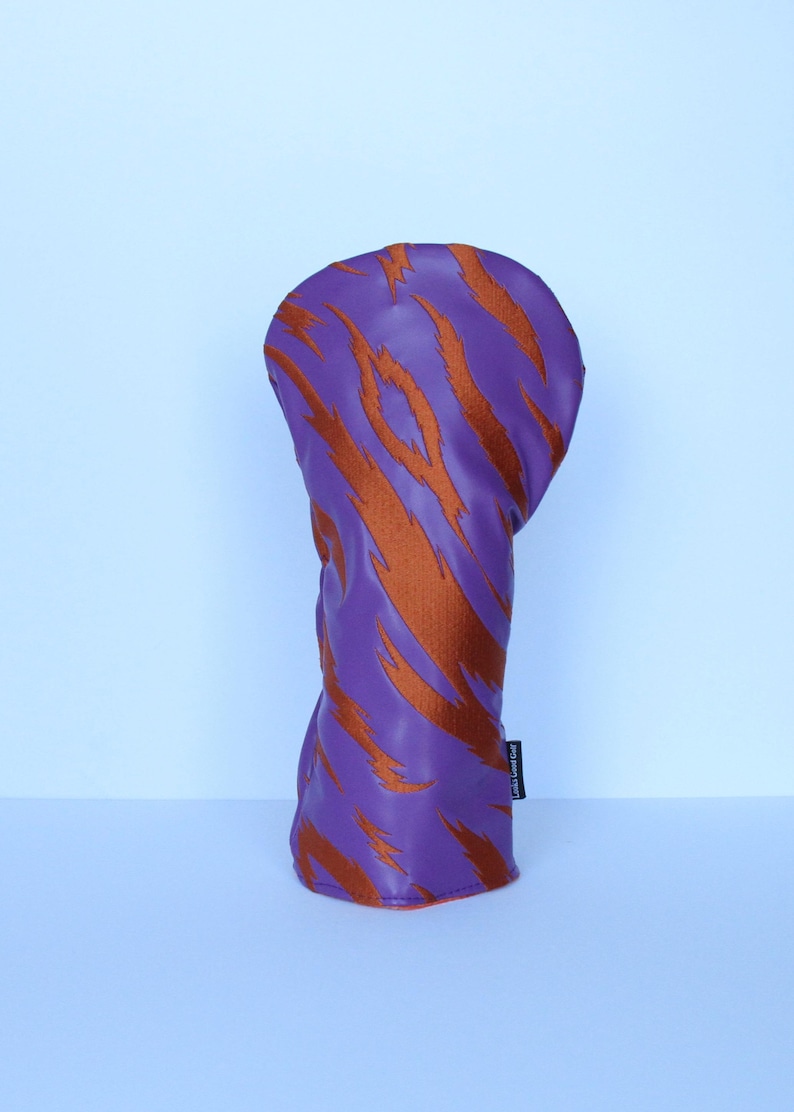 May include: Purple golf club head cover with orange tiger stripe design. The cover is made of a smooth material and has a rounded top. The cover is designed to protect the golf club head.