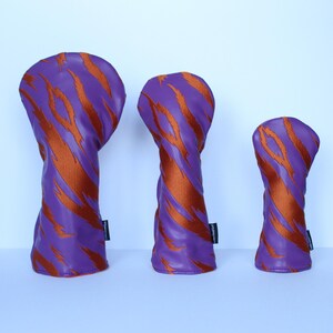 May include: Three golf club head covers in a row. The covers are purple with orange tiger stripe patterns. Each cover has a black tag on the side. The covers are in different sizes.