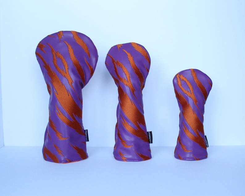 May include: Three golf club head covers in a row. The covers are purple with orange tiger stripe patterns. Each cover has a black tag on the side. The covers are in different sizes.