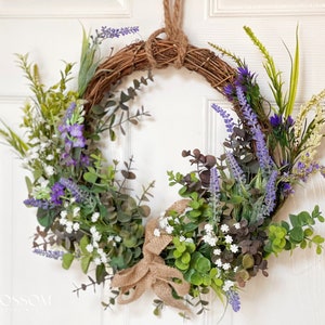 May include: A wreath made of woven twigs with a hessian bow. The wreath is decorated with purple lavender flowers, white flowers, and green leaves.
