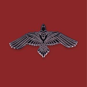 Peut inclure: Épingle en métal représentant un corbeau aux ailes déployées. Le corps et les ailes sont noirs, avec des détails argentés. La poitrine du corbeau présente un motif de nœud celtique. Fond rouge uni.